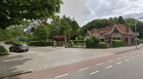 Een straatbeeld met een toegangspoort tot een park, omgeven door groen. Links is een geparkeerde auto te zien en rechts een karakteristiek bakstenen huis met een rood dak. De straat heeft een fietspad en trottoir. Op de achtergrond zijn hoge bomen en een bewolkte lucht zichtbaar.